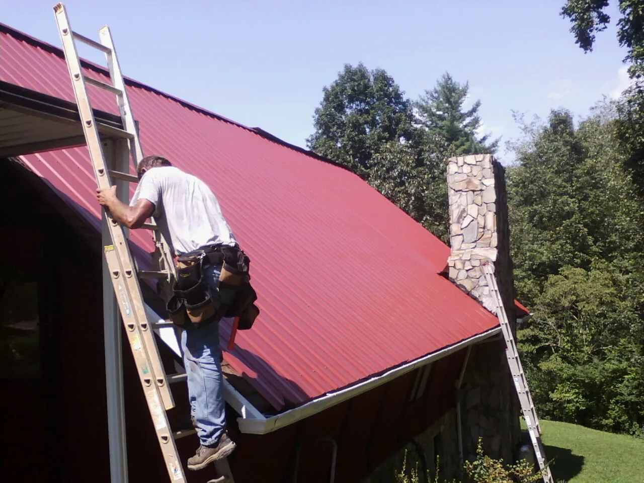 Roofer on a ladder beside a red metal roof and stone chimney — Soffit & Fascia Repair by Roofing Brothers in Tullahoma